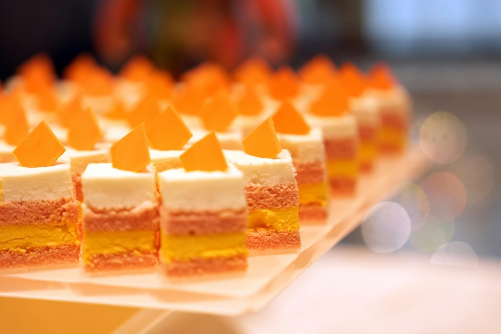 Row of neatly arranged colorful layered cakes with orange triangular toppings on a white platter.