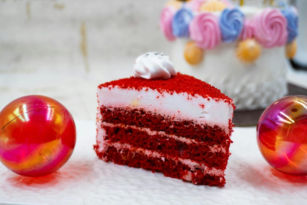 A vibrant red velvet cake slice with frosting, surrounded by festive colorful decorations.